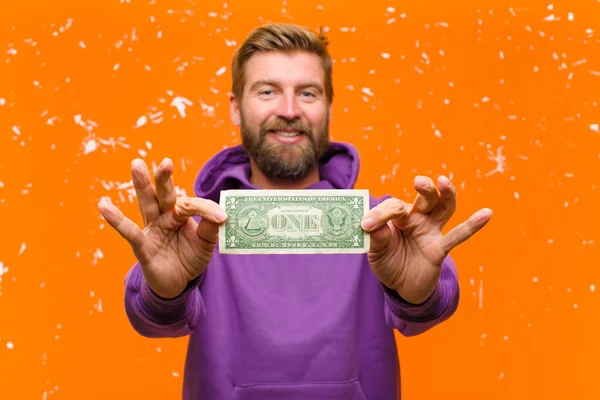 年轻的金发男子 戴着美元钞票或钞票 头戴紫色的帽衫 靠着残破的橙色墙壁 — 图库照片