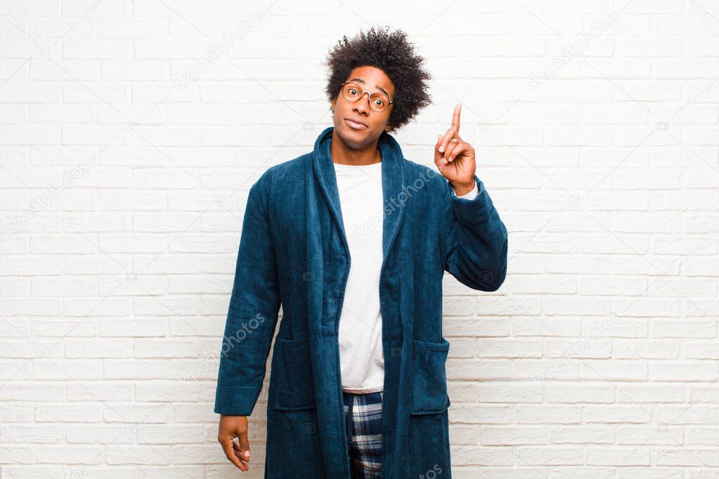 young black man wearing pajamas with gown feeling like a genius holding finger proudly up in the air after realizing a great idea, saying eureka against brick wall