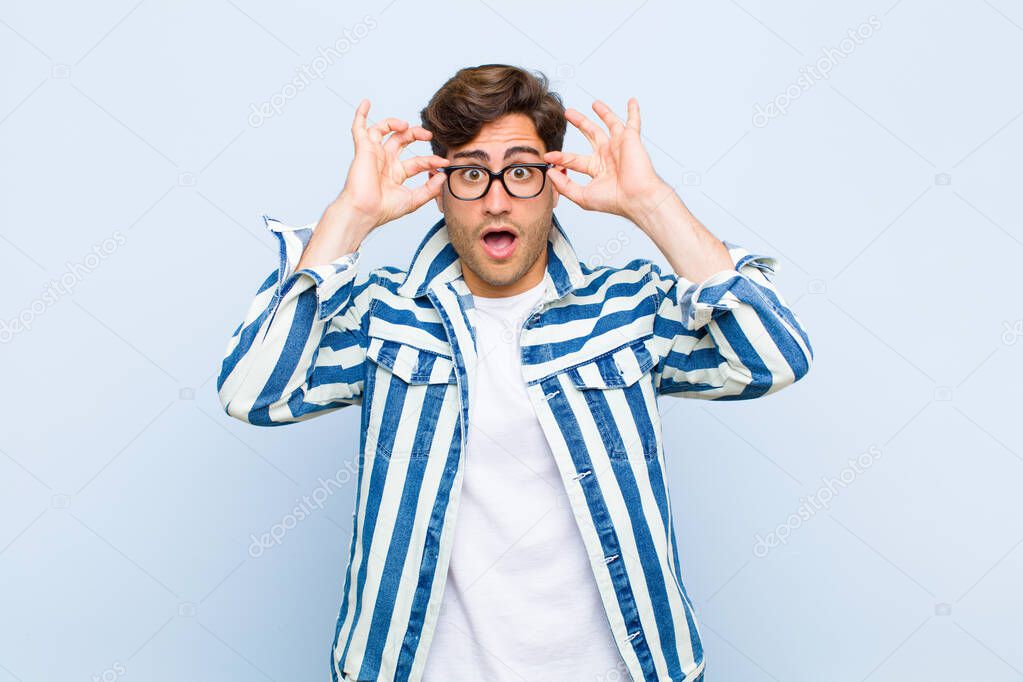 young handsome man feeling shocked, amazed and surprised, holding glasses with astonished, disbelieving look against blue background