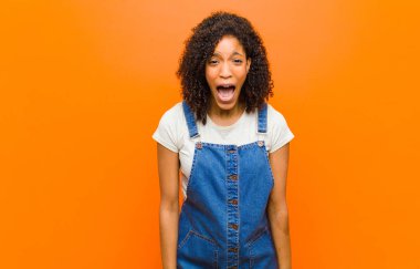 Genç, güzel, siyah bir kadın agresif bir şekilde bağırıyor. Kızgın, sinirli ya da sinirli görünüyor. Turuncu duvara karşı hayır diye bağırıyor.