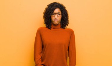 young pretty black woman feeling puzzled and confused, with a dumb, stunned expression looking at something unexpected against orange wall