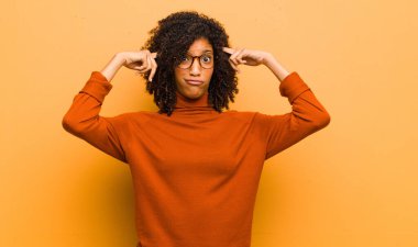 Ciddi ve konsantre görünümlü genç, güzel siyahi kadın, turuncu duvara karşı zorlu bir sorun düşünüyor ve beyin fırtınası yapıyor.