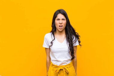 Şok olmuş, kızgın, hayal kırıklığına uğramış, ağzı açık ve turuncu duvara karşı öfkeli görünen genç Latin bir kadın.