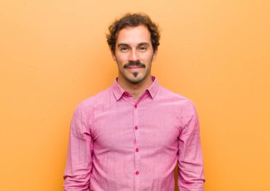 Genç yakışıklı adam pozitif ve kendinden emin gülümsüyor. Tatmin olmuş, arkadaş canlısı ve turuncu duvarın karşısında mutlu görünüyor.