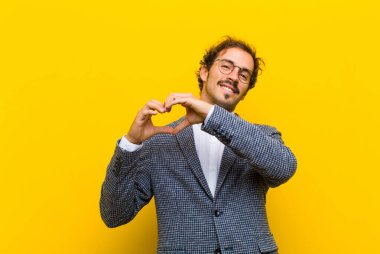 Genç yakışıklı adam gülümsüyor ve mutlu, sevimli, romantik ve aşık, iki elini turuncu duvara dayayıp kalp şekli veriyor.