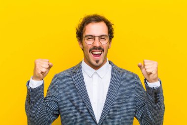 Genç yakışıklı adam mutlu, şaşırmış ve gururlu hissediyor, bağırıyor ve başarıyı kutluyor turuncu duvarda kocaman bir gülümsemeyle.