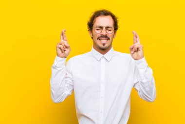 Genç yakışıklı adam gülümsüyor ve endişeyle iki parmağını çapraz yapıyor. Endişeli ve turuncu duvara karşı iyi şanslar diliyor ya da umut ediyor.