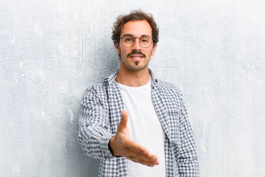 Genç yakışıklı adam gülümsüyor, mutlu, kendinden emin ve arkadaş canlısı görünüyor. Anlaşmayı bağlamak için el sıkışmayı teklif ediyor. Düz duvara karşı izole edilmiş bir şekilde işbirliği yapıyor.
