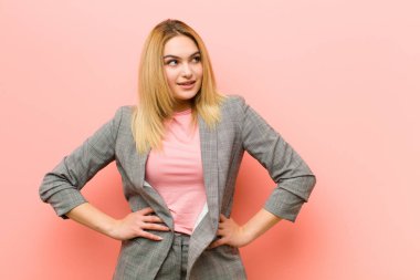 Genç, güzel, sarışın kadın mutlu, neşeli ve kendinden emin görünüyor gururla gülümsüyor ve iki elini de düz duvara yaslamış kalçaları üzerinde görmek istiyor.