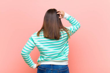 Genç, güzel, İspanyol bir kadın kendini cahil ve kafası karışık hissediyor, bir çözüm düşünüyor, eli kalçasında, diğeri kafasında, pembe duvarın arkasında dikiz aynasıyla.