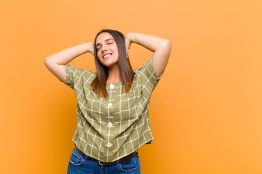 Genç ve güzel bir kadın gülümsüyor ve rahatlıyor, tatmin oluyor ve kaygısız, gülüyor ve turuncu duvarda izole bir şekilde dinleniyor.