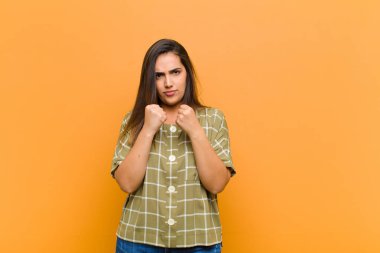 Genç, güzel bir kadın kendinden emin, sinirli, güçlü ve agresif görünüyor. Yumrukları boks pozisyonunda dövüşmeye hazır turuncu duvara karşı izole.