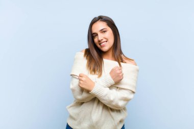 Genç ve güzel bir kadın sevinçle gülümsüyor ve kutluyor, yumruklarını sımsıkı kavrayıp kollarını kavuşturuyor, mutlu ve pozitif hissediyor mavi arka plana karşı.