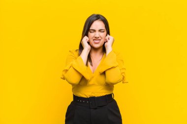 Genç, güzel bir kadın kızgın, stresli ve sinirli görünüyor, her iki kulağı da sağır edici bir ses, ses veya yüksek sesle kapatıyor turuncu duvarda izole edilmiş bir müzik