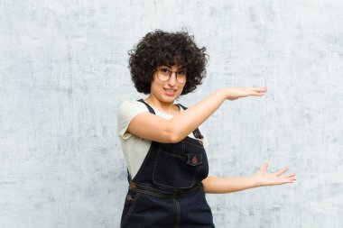 Genç, güzel, afro bir kadın iki eli yanda bir objeyi tutuyor fotokopi alanında, bir nesneyi gösteriyor, sunuyor ya da reklamını yapıyor.