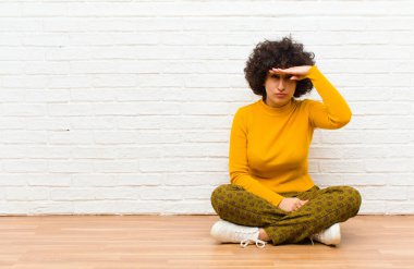 Genç ve güzel afro kadın şaşkınlık ve hayret içinde bakıyor, el üstünde uzak bakıyor, izliyor ya da arıyor yerde oturuyor