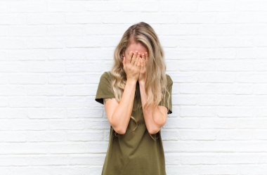 Genç sarışın kadın üzgün, hayal kırıklığına uğramış, gergin ve depresif, iki eliyle yüzünü örten, tuğla duvar arkasında ağlayan