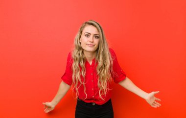 Genç sarışın kadın, kafası karışık ve bihaber, hiçbir fikri yok, kırmızı duvara karşı aptal ya da aptal bir bakışla şaşırmış.