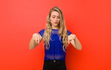 Genç sarışın kadın, ağzı açık, iki eliyle aşağıya bakıyor, şaşkın, şaşkın ve şaşırmış görünüyor kırmızı duvara karşı