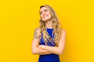 Genç sarışın kadın kendini mutlu, gururlu ve umutlu hissediyor, merak ediyor veya düşünüyor, sarı duvara yaslanmış çapraz kollarla boşluğu doldurmaya çalışıyor.