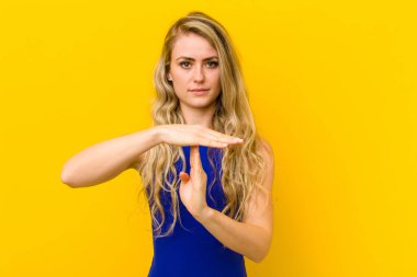 Genç sarışın kadın ciddi, sert, kızgın ve hoşnutsuz görünüyor. Sarı duvara karşı mola işareti yapıyor.