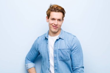 Genç kızıl saçlı adam mutlu bir şekilde gülümsüyor, eli kalçasında ve kendinden emin, pozitif, gururlu ve dost canlısı bir tavırla mavi duvara karşı.