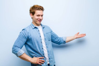 Genç kızıl saçlı adam kendini mutlu ve neşeli hissediyor, gülümsüyor ve seni selamlıyor, seni mavi duvara karşı dostça bir jestle içeri davet ediyor.