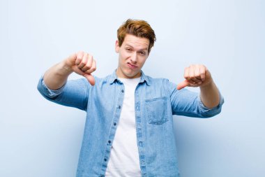 Genç kızıl saçlı adam üzgün, hayal kırıklığına uğramış ya da kızgın görünüyor, anlaşmazlık içinde başparmaklarını gösteriyor, mavi duvara karşı hayal kırıklığına uğramış hissediyor.