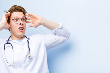 Genç, kızıl saçlı, ağzı açık genç bir doktor korkunç bir hata yüzünden dehşete düşmüş ve şok olmuş görünüyor. Mavi duvara karşı ellerini kaldırıyor.