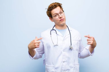 Kötü tavırlı genç kızıl saçlı doktor gururlu ve agresif görünüyor, yukarı bakıyor ya da mavi duvara yaslanmış ellerle dalga geçiyor.