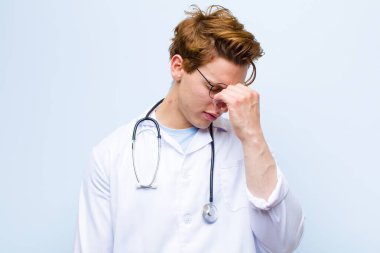 Genç kızıl kafalı doktor stresli, mutsuz ve hayal kırıklığına uğramış, alnına dokunmuş ve mavi duvara karşı şiddetli baş ağrısı çeken.