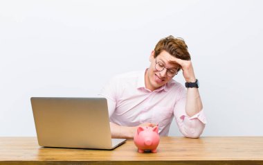 Genç kızıl saçlı işadamı masasında domuzcuk kumbarasıyla çalışıyor.