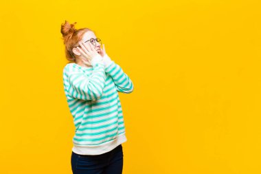 Genç kızıl saçlı kadın mutlu, heyecanlı ve şaşırmış hissediyor, yüzünü turuncu duvara dayamış, yan tarafa bakıyor.