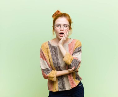 Genç ve güzel kızıl saçlı kadın şok ve inançsızlık içinde, elleri yanak ve kolları çapraz, sersemlemiş ve şaşırmış hissediyor yeşil duvara karşı
