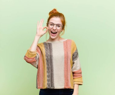 Genç ve güzel kızıl saçlı kadın kendini başarılı ve tatmin olmuş hissediyor, ağzı açık gülümsüyor, yeşil duvara karşı elle işaret veriyor.