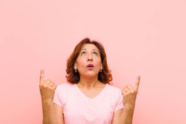 Orta yaşlı bir kadın korkuyla ve ağzı açık bir şekilde pembe duvarlara karşı şaşkın ve şaşkın bir bakışla yukarıya bakıyor.