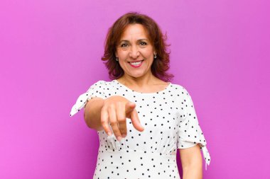 Orta yaşlı bir kadın kameraya bakıyor. Tatmin olmuş, kendine güvenen, arkadaş canlısı bir gülümsemeyle. Seni mor duvara karşı seçiyor.