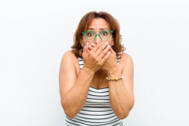 Orta yaşlı, güzel bir kadın şok olmuş, şaşırmış bir ifadeyle ağzını kapatıyor. Sır saklıyor ya da beyaz duvara karşı 