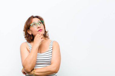 Orta yaşlı güzel bir kadın düşünceli, meraklı ya da hayal kuruyor, hayal kuruyor ve beyaz duvara karşı boşluğu kopyalamak için yukarı bakıyor.