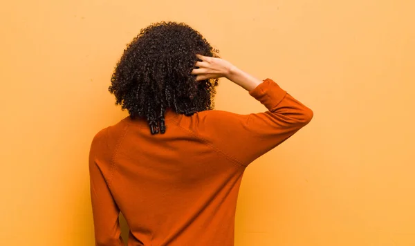 young pretty black woman thinking or doubting, scratching head, feeling puzzled and confused, back or rear view against orange wall