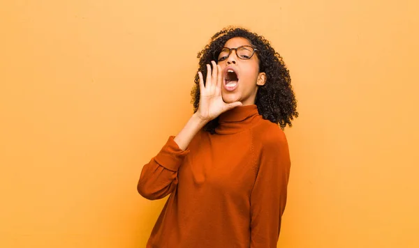 young pretty black woman yelling loudly and angrily to copy space on the side, with hand next to mouth against orange wall