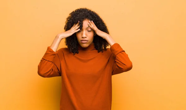 young pretty black woman looking stressed and frustrated, working under pressure with a headache and troubled with problems against orange wall