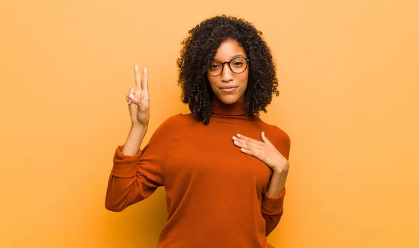 young pretty black woman looking happy, confident and trustworthy, smiling and showing victory sign, with a positive attitude against orange wall