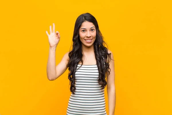 年轻漂亮的拉丁女子感到快乐 放松和满意 以好的姿态表示赞同 靠在橙色的墙上微笑 — 图库照片