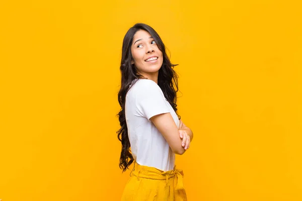 young pretty latin woman smiling gleefully, feeling happy, satisfied and relaxed, with crossed arms and looking to the side against orange wall