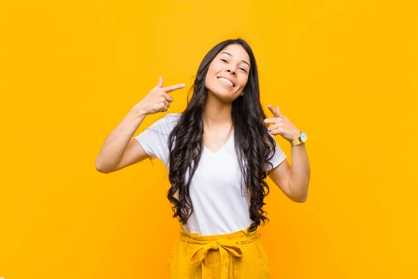 young pretty latin woman smiling confidently pointing to own broad smile, positive, relaxed, satisfied attitude against orange wall