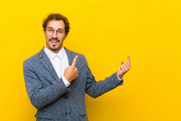 young handsome man smiling happily and pointing to side and upwards with both hands showing object in copy space against orange wall