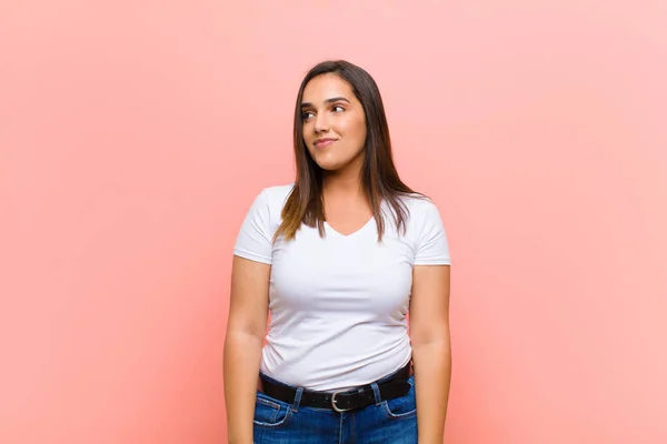 young pretty hispanic woman wondering, thinking happy thoughts and ideas, daydreaming, looking to copy space on side against pink wall