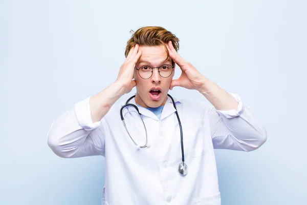 young red head doctor raising hands to head, open-mouthed, feeling extremely lucky, surprised, excited and happy against blue wall