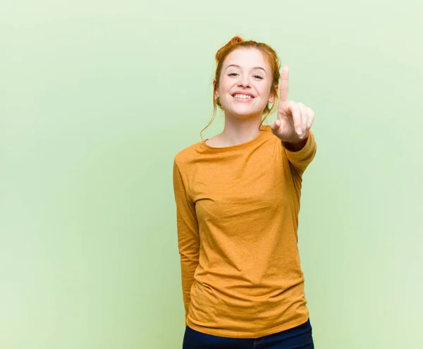 若いかなり赤い頭の女性は笑顔で友好的に見えます前方に手で1番または1番を示し緑の壁に対してカウントダウン — ストック写真